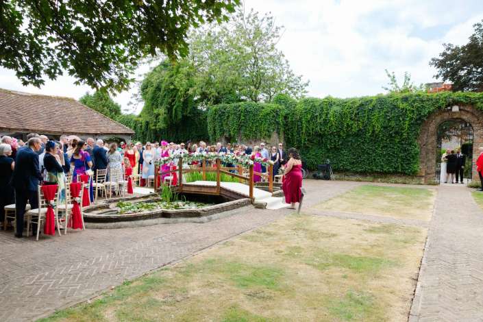 outdoor wedding ceremony with guests Outdoor wedding ceremony in an ivy-covered courtyard with a flower-decorated wooden bridge over a pond and guests in formal attire.