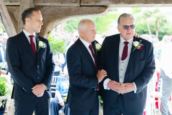 three groomsmen at wedding Three men in dark suits with red ties and white rose boutonnieres stand together at an outdoor wedding.