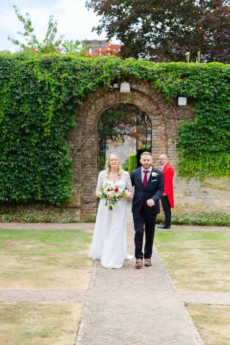bride and groom exit garden gate Bride in a white wedding gown and groom in a dark suit walk down a cobblestone path beneath an ivy-covered brick archway.