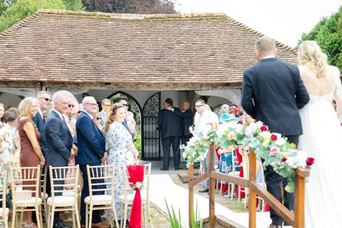 rustic outdoor wedding ceremony Outdoor wedding ceremony with guests in formal attire gathered along a white aisle as the bride and groom stand at the altar beside a rustic thatched-roof building.