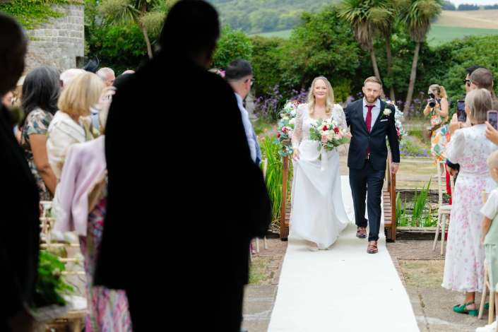 bride and groom walking aisle Bride in a white wedding gown holding a bouquet walks down an outdoor aisle beside a groom in a dark suit, with guests and photographers on either side.