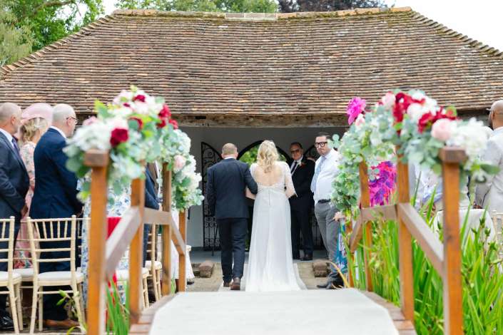 rustic outdoor wedding aisle Bride in a white wedding dress and her groom walk down an outdoor aisle flanked by colorful floral arrangements, with guests standing on both sides.