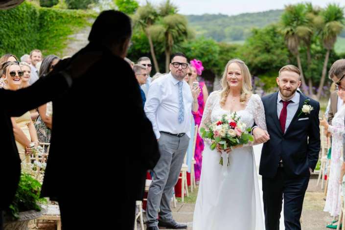 bride and groom entrance Bride in a lace wedding dress carrying a bouquet walks with her groom down an outdoor aisle as guests smile and watch.
