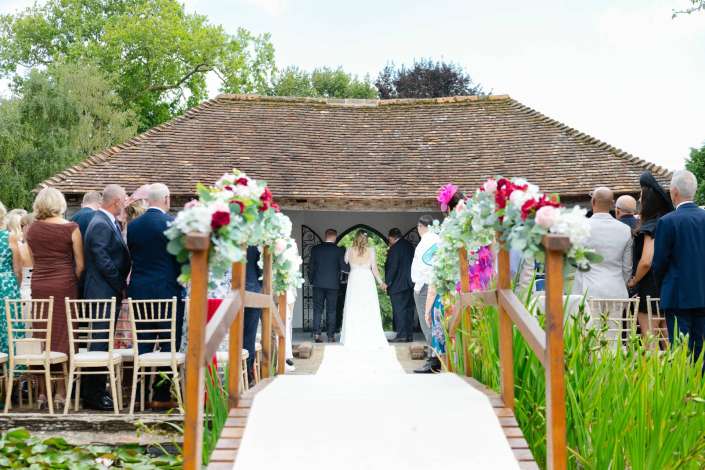 rustic outdoor wedding ceremony Outdoor wedding ceremony with guests gathered along a flower-decorated white aisle toward a bride and groom under a rustic tile-roofed shelter.