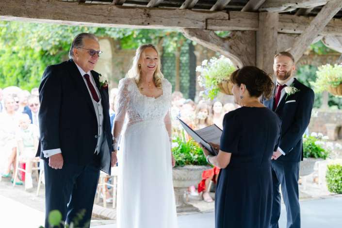outdoor wedding under wooden pergola Bride and groom stand under a rustic wooden shelter during an outdoor wedding, with an officiant reading from a folder and guests visible in the background.