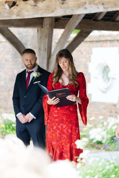 couple reading wedding vows A man in a navy suit with a boutonniere stands beside a woman in a red dress who is reading from a dark book at an outdoor ceremony.