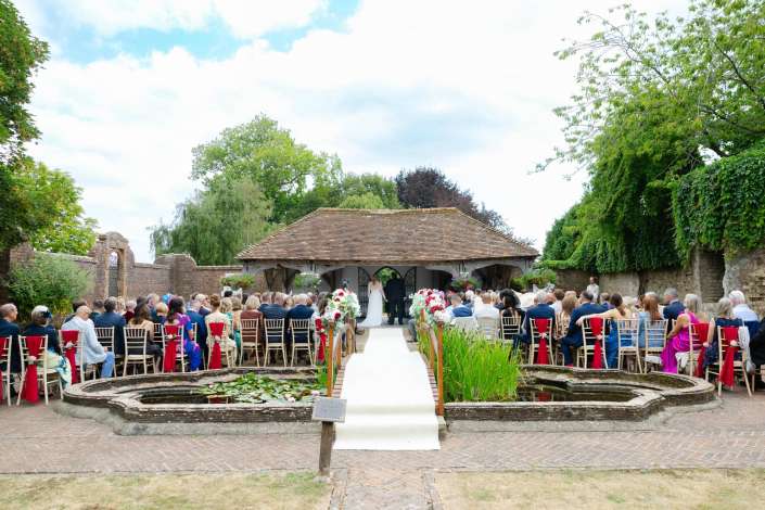 outdoor garden wedding ceremony Outdoor wedding ceremony in a garden with a thatched pavilion, rows of chairs along a white aisle, and a circular fountain in the foreground.