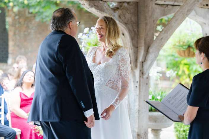 bride and groom exchange vows Bride in a white lace gown smiles at her groom during an outdoor wedding ceremony beneath a wooden pergola.
