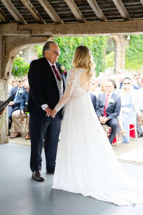 bride and father hold hands Bride in a lace long-sleeve wedding gown with an open back holds hands with an older man in a suit under a rustic wooden arch at an outdoor wedding.