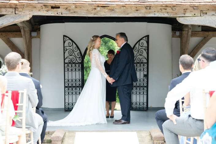 bride and groom exchange vows Bride in a lace wedding gown and groom in a dark suit hold hands and smile at each other during a rustic outdoor wedding ceremony framed by decorative wrought-iron gates.