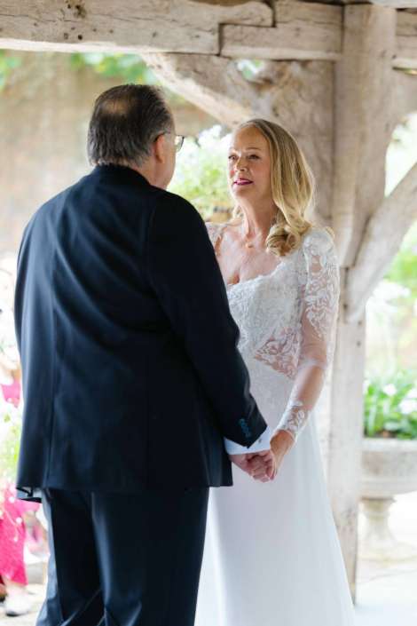 bride and groom holding hands Bride in a lace wedding dress and her partner in a dark suit hold hands and look at each other under a rustic wooden shelter.