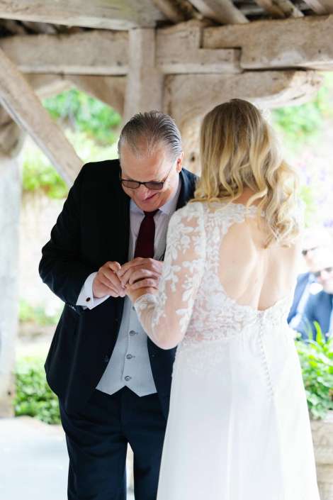 father and bride ring exchange Groom in a dark suit and vest placing a ring on the bride's finger during an outdoor wedding under a rustic stone structure.
