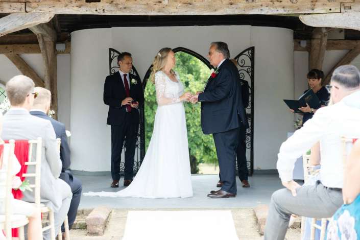 bride and groom exchange rings Bride in a white wedding dress exchanges rings with a suited man during an outdoor ceremony under a rustic wooden shelter, with guests seated and an officiant reading.