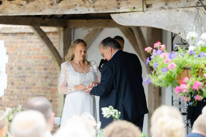 bride and groom exchange rings Bride in a lace wedding dress and groom in a dark suit exchange rings under a rustic wooden arch with colorful flowers and guests nearby.