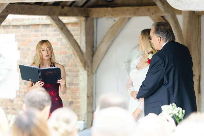officiant reading wedding vows Bride in a white lace wedding dress and groom in a dark suit stand with an officiant reading from a large binder under rustic wooden beams.