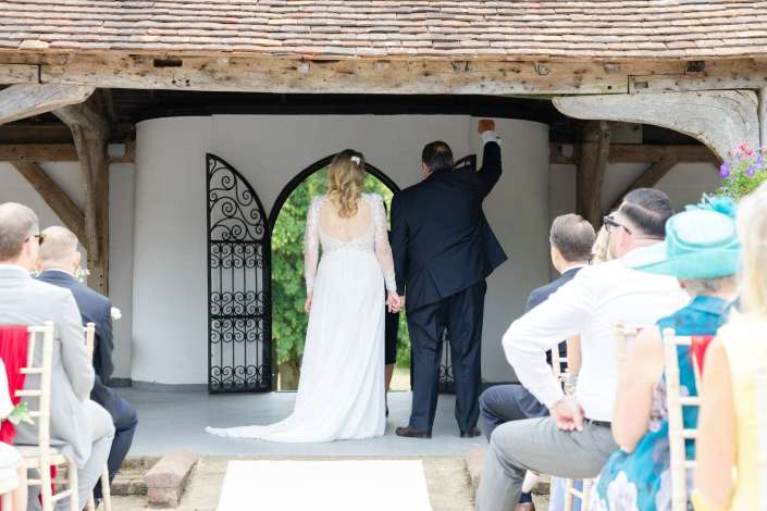 rustic wedding arch entrance Bride in a lace-back wedding gown and groom in a dark suit stand with their backs to the camera at an outdoor rustic wedding, in front of an arched wrought-iron gate.