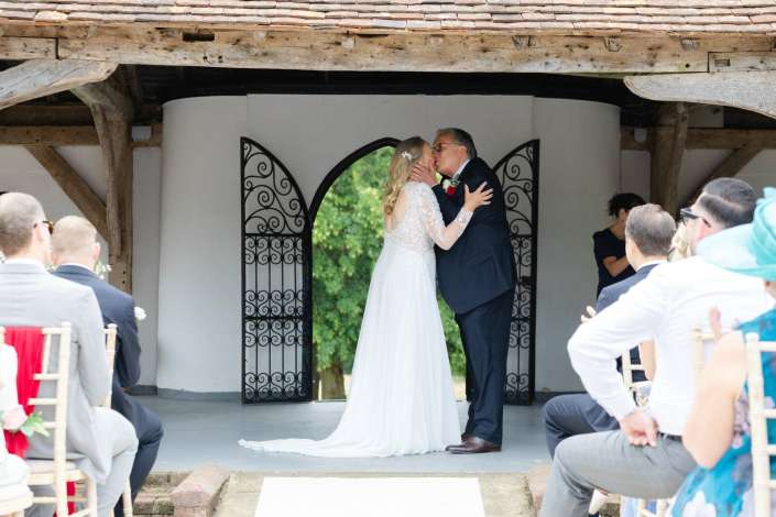 romantic wedding kiss under arch Bride in a lace wedding dress and groom in a dark suit kiss at a rustic outdoor altar as guests look on.