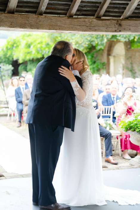 bride and father kiss Bride in a white lace wedding dress kisses her father during the ceremony, with guests watching in the background.