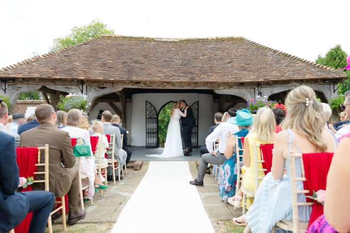 romantic outdoor wedding ceremony Bride and groom kiss at the end of a white aisle in a rustic outdoor wedding under a thatched-roof shelter, surrounded by seated guests.