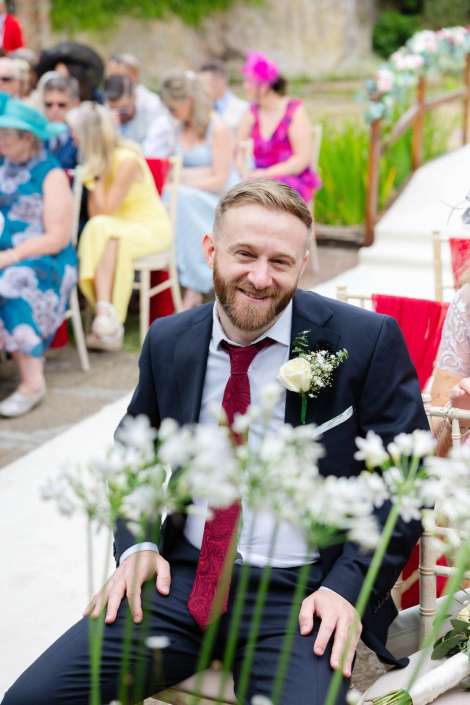 smiling groom at garden wedding A smiling man in a navy suit with a white rose boutonniere sits at an outdoor wedding as guests in colorful dresses are seated behind him.