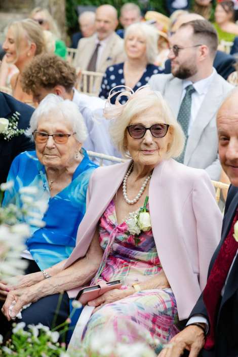 elegant elderly woman at wedding Two elderly women seated at an outdoor wedding; the front woman wears a pink blazer, floral dress, pearl necklace and sunglasses.
