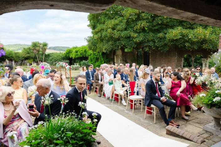 garden wedding ceremony guests Outdoor wedding ceremony in a lush garden with guests seated along a white aisle under a large leafy tree beside a brick wall.