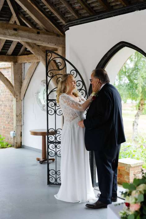 bride embraces her father Bride in a white lace gown gently touches the face of a man in a dark suit beneath an ornate iron arch inside a rustic venue.