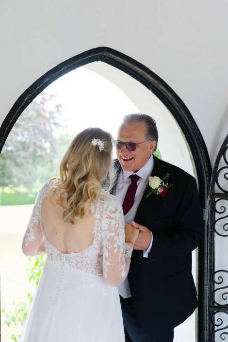 joyful father-daughter moment Bride in a lace wedding dress and an older man in a dark suit smile while holding hands beneath a black arched doorway.