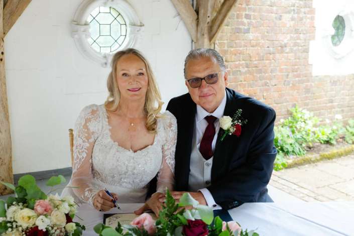 bride and groom signing wedding documents A bride in a lace wedding dress signs a document at a table adorned with flowers, while her groom in a dark suit sits beside her in a rustic brick venue.
