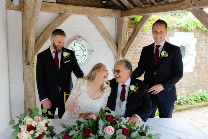 bride and groom with groomsmen Bride in a lace wedding dress sits with an elderly couple at a flower-decorated table, flanked by two smiling groomsmen in suits under a rustic wooden shelter.