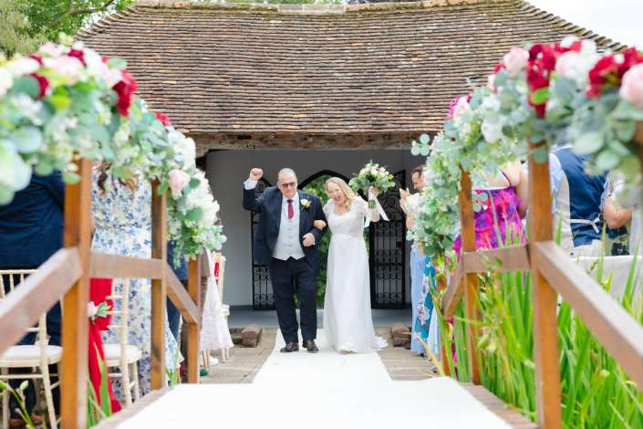 bride and father walk down aisle Bride in a white wedding gown walks down a flower-decorated outdoor aisle with her father in a suit as guests applaud along the sides.