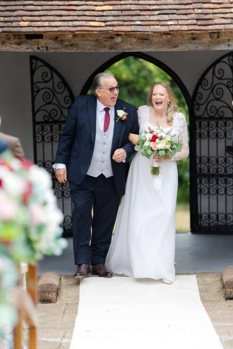 bride and father walking down aisle Bride in a white wedding dress walks arm-in-arm with her father in a dark suit, both laughing, holding a bouquet.