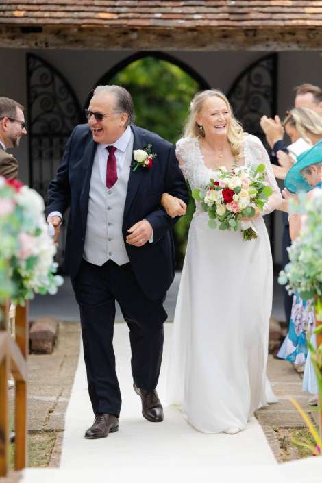bride walks down the aisle Bride in a white lace wedding dress walks arm-in-arm with a man in a dark suit down an outdoor aisle, smiling, holding a bouquet.