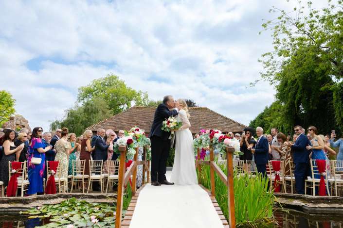 outdoor wedding kiss on bridge Bride and groom kiss on a white aisle during an outdoor wedding, with guests on both sides and colorful floral arrangements along a wooden bridge over a pond.