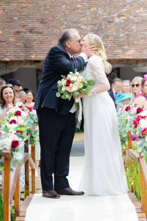 father kissing bride at wedding Bride in a white gown kisses a man in a suit on the wedding aisle, holding a pink and white bouquet.