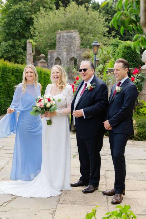 joyful outdoor wedding portrait Bride in a white lace gown holding a bouquet stands with a bridesmaid in a pale blue dress and two men in dark suits outdoors.