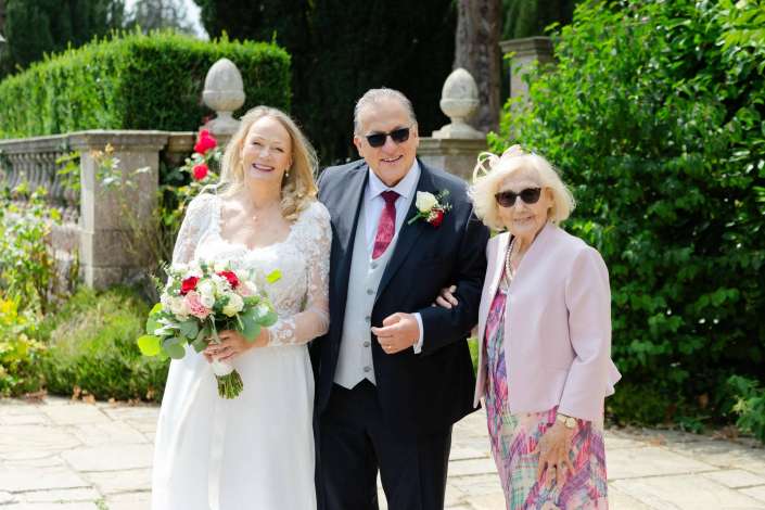 bride with her parents Smiling bride in a white wedding dress holds a bouquet, standing with a man in a suit and an older woman in a pink blazer in a garden.