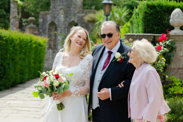joyful outdoor wedding portrait Bride in a lace wedding dress holds a bouquet while standing with a man in a suit and an elderly woman in a pink blazer in a sunny garden.