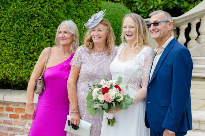 wedding party portrait outdoors Four adults posing outdoors on stone steps, including a bride in a white lace gown holding a bouquet, flanked by two women in colorful dresses and a man in a blue suit against a green hedge backdrop.