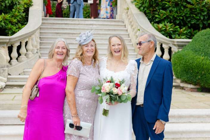 bride with family on stairs Smiling bride in a white wedding dress holds a bouquet while posing with two women and a man in formal attire on a stone staircase with greenery.