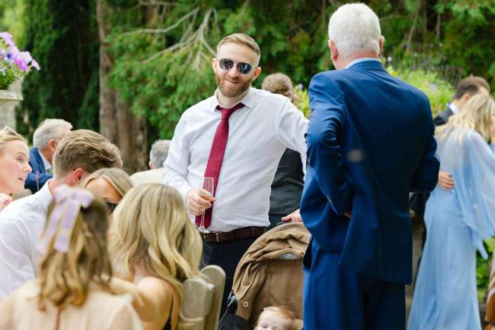 smiling guests at outdoor wedding A smiling man in a white shirt, red tie, and sunglasses chats with a man in a blue suit at an outdoor wedding reception.