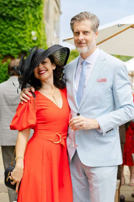 elegant couple at garden party A couple posing at an outdoor garden party, the woman in a bright red dress with a black wide-brim hat and the man in a light blue suit holding a champagne flute.
