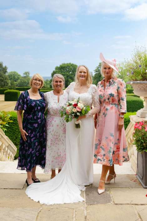 bridal party in floral dresses Bride in a white wedding gown with a long train holding a bouquet, flanked by three women in pastel floral dresses on a sunny garden terrace.