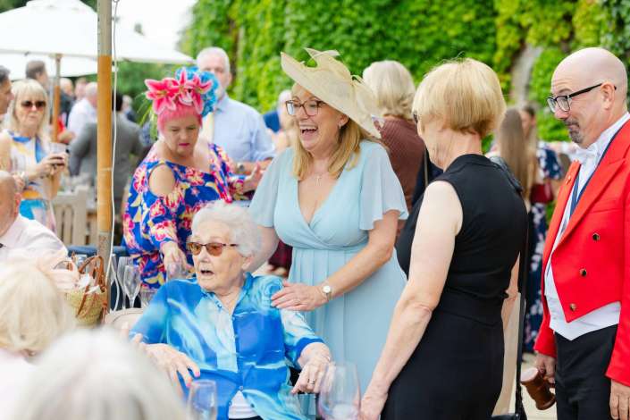 joyful garden party guests Group of elegantly dressed people at an outdoor garden party, featuring a smiling woman in a pale blue dress and large beige hat.