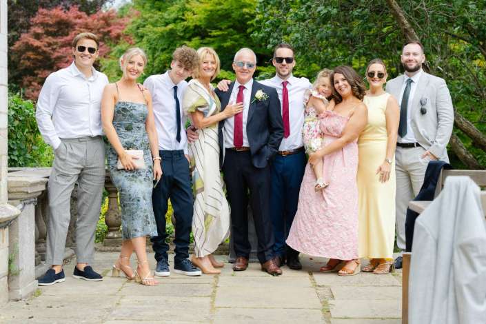 group wedding portrait outdoors A wedding party stands together outdoors in a green garden, dressed in formal attire.