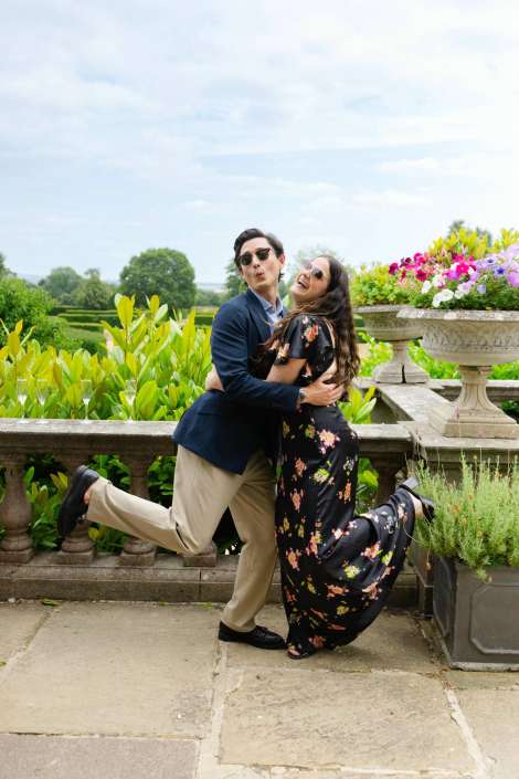 joyful garden embrace A couple embraces and smiles on a stone terrace, the man in a navy blazer and beige trousers and the woman in a black floral dress, with colorful flower planters in the background.