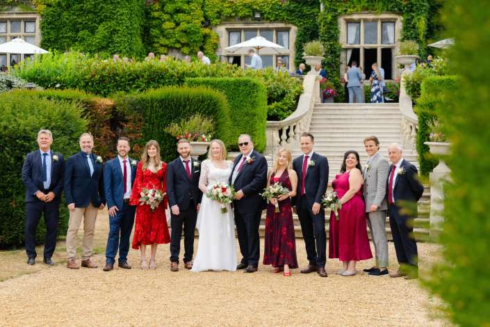 outdoor wedding party Wedding party posing outdoors on a gravel path in front of an ivy-covered stone mansion, with the bride and groom center and bridesmaids in red dresses and groomsmen in suits.