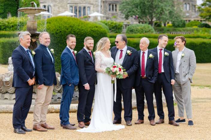wedding party at fountain Bride in a lace white wedding dress holds a bouquet while standing with her groom and groomsmen in suits outdoors by a fountain and manicured garden.