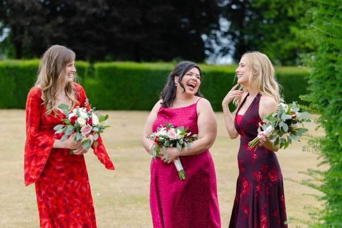 bridesmaids laughing outdoors Three women in red and pink dresses stand outdoors, smiling and holding bouquets of flowers.