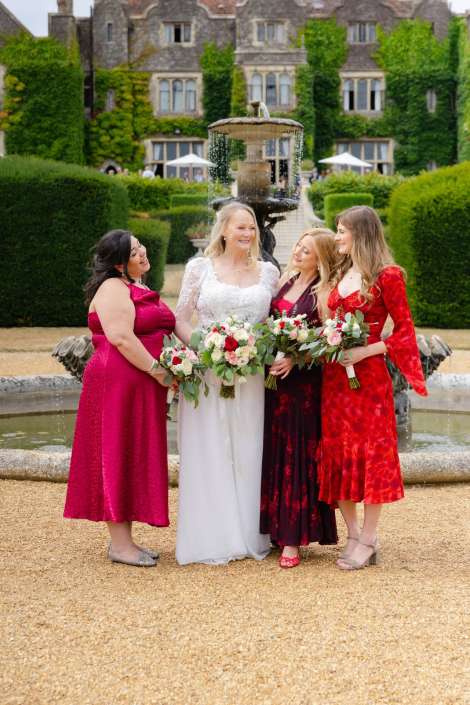 bridal party by fountain A bride in a white wedding dress stands with three bridesmaids in pink, burgundy, and red dresses, all holding bouquets in front of a fountain and formal garden.